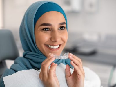 Orthodontics Concept. Smiling Muslim Woman In Hijab Holding Invisalign Or Invisible Braces While Sitting At Dentists Chair In Clinic, Islamic Female Using Clear Dental Aligner For Teeth Correction